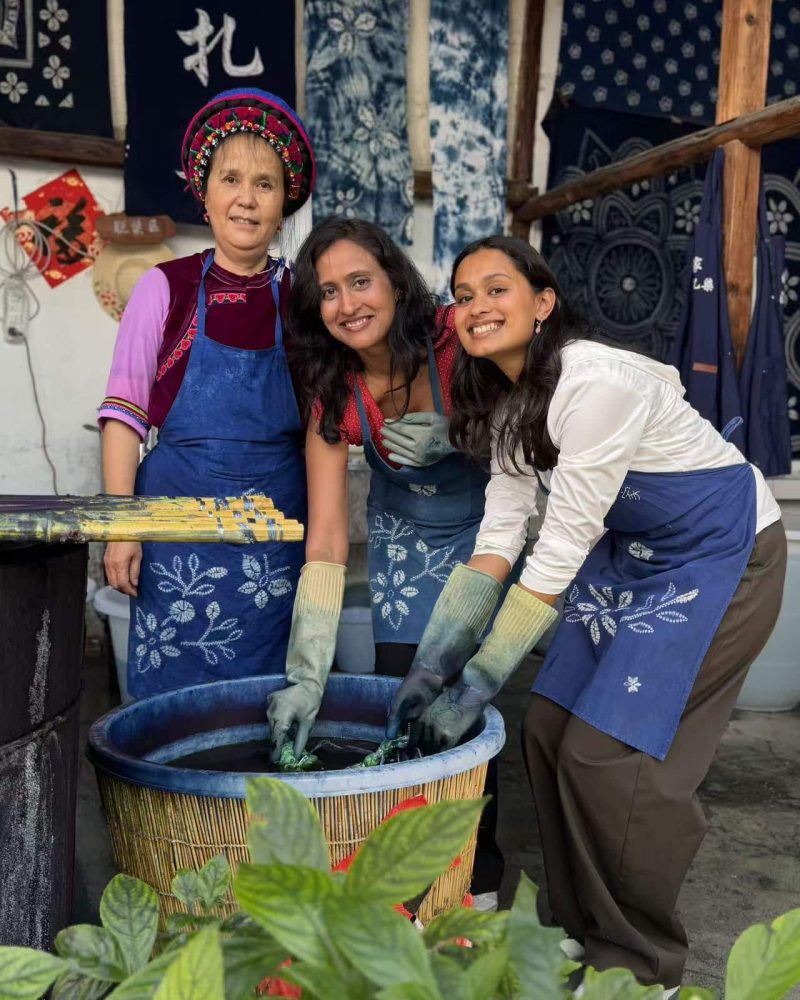 Three participants in blue aprons at an indigo tie-dye workshop, with patterned fabrics hanging behind them