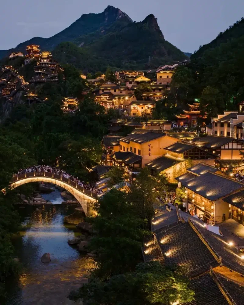 Illuminated hillside buildings and an arched bridge follow a stream beneath the mountains at Wangxian Valley.