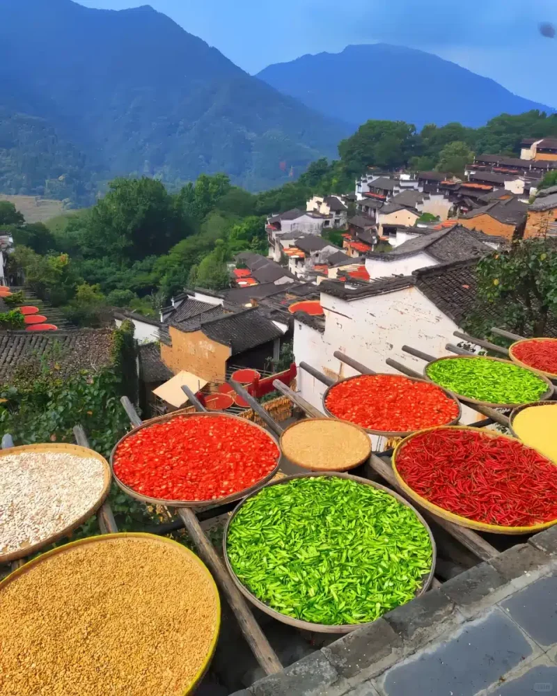 Colorful round drying trays fill the foreground above tiled rooftops in Huangling village beneath wooded hills.
