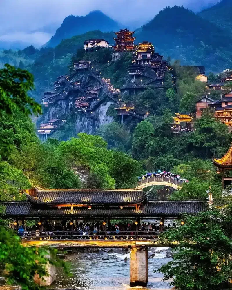 Illuminated houses cling to a misty cliff above an arched stone bridge and a covered wooden bridge in Wangxian Valley.