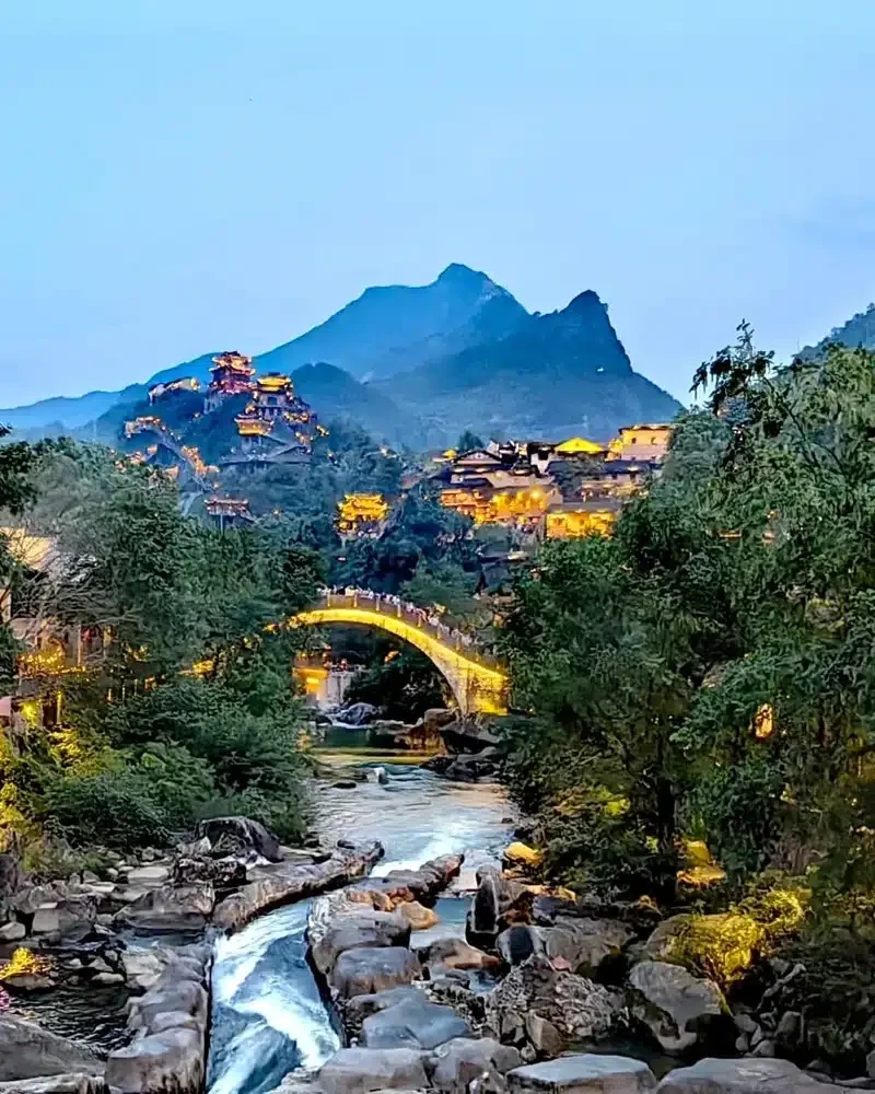 A rocky stream runs below an illuminated stone arch bridge, with hillside buildings and mountain peaks behind it in Wangxian Valley.