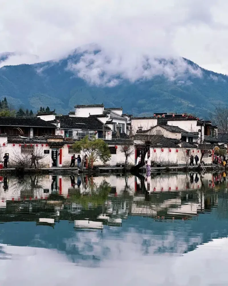 White-walled houses and dark tiled roofs reflect in still water beneath misty hills at Hongcun village.