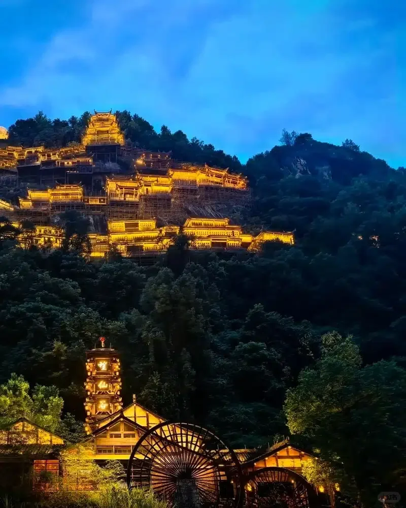 Illuminated cliffside buildings rise above a water wheel and a golden Wangxian Valley sign at night.