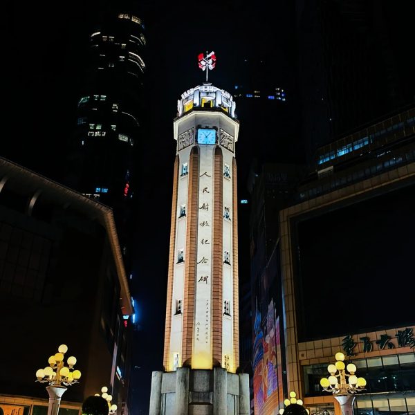 Jiefangbei Monument illuminated at night in downtown Chongqing, surrounded by skyscrapers and neon lights.
