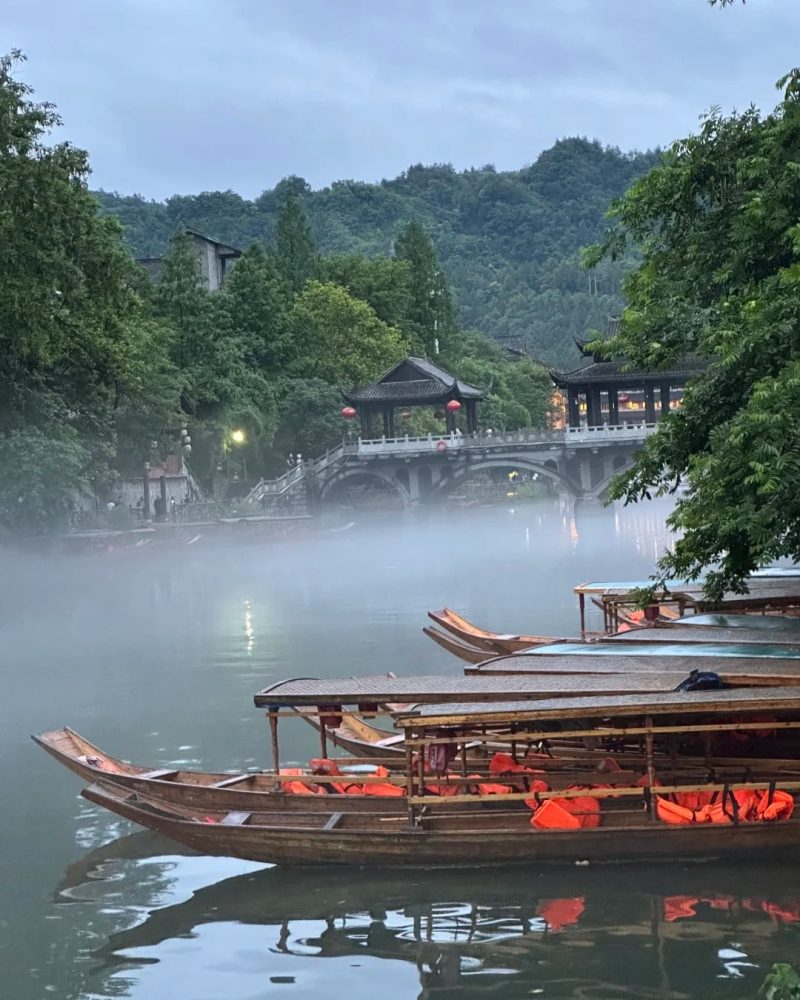 Boats on the river in Phoenix Ancient Town, China, with mist, trees, and an old stone bridge